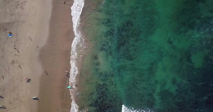 Timelapse. High aerial top view of tiny surfers on sand beach. Small toy figures go into water. Sea waves on beautiful beach aerial view drone shot. Bird's eye view of ocean waves crashing at beach.