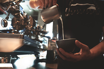 barista man pouring whipped milk from frothing pitcher in cup with coffee standing in front of professional coffee machine in cafe