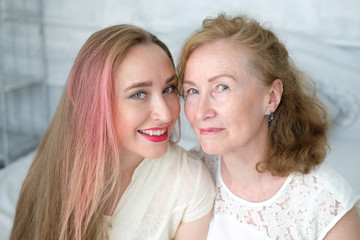 Beautiful old woman with her adult daughter in the bedroom. The woman has blond curly hair, kind eyes and smile. Daughter with long hair and bright lips. Beautiful aging, love, motherhood