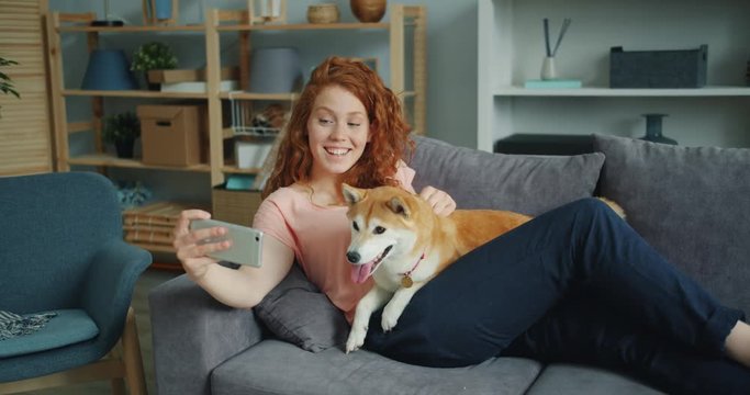 Happy young girl is taking silfie with lovable dog using smartphone camera having fun sitting on couch holding device. Youth and domestic animals concept.