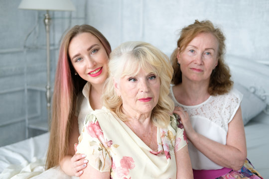 Three generations of women sitting on the bed in the bedroom. An adult daughter, her mother and grandmother. Beautiful girl with long hair. The old woman has gray curls, chiffon dress with flowers.