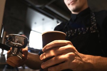 barista man in apron holding hot coffee drink in paper cup in one hand and holder in another. standing close to professional coffee machine in cafe close up