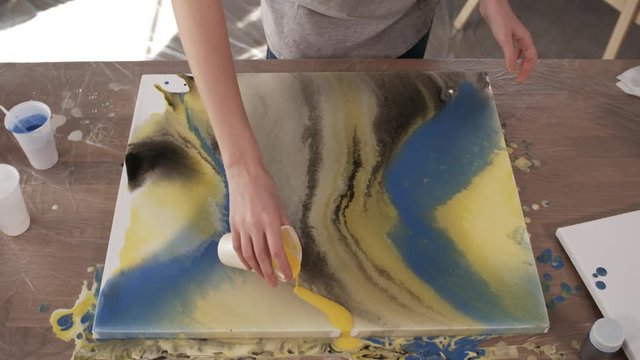 Top view of female painter pouring yellow acrylic paint over canvas and tilting it while creating abstract painting at her workplace in art studio