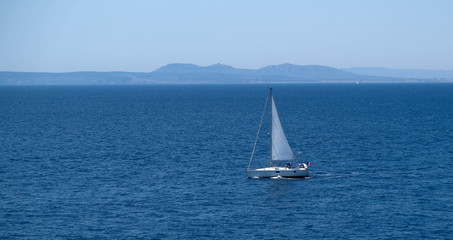 Fototapeta premium Small sailboat at the sea. Sailing near the shore in the mediterranean sea. Amazing sunny day, calm blue water. Sail race in the coastal bay. Boat in the sea. Happy vacation. White sail.