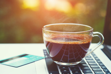 transparent cup of hot black coffee, credit card and open laptop on wooden table, sunny green blurred summer background, copy space