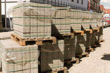 Plastersteine aus Beton auf Holzpaletten in Reihe und übereinander gestapelt, mit Zurrband gesichert und abgedeckt mit Plastikfolie als Regenschutz