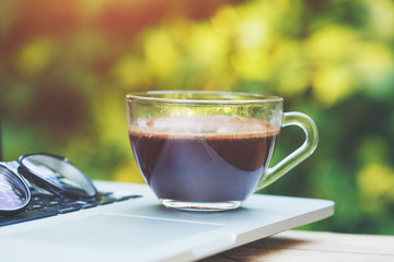 transparent cup of coffee, glasses and open laptop on wooden table, green blurred background, hot sunny summer day