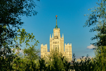 View of Stalin skyscraper on Kotelnicheskaya Embankment through foliage of trees from Zaryadye Park. Stalin skyscraper in rays of evening sun against blue cloudless sky Moscow, Russia, June, 2019: 