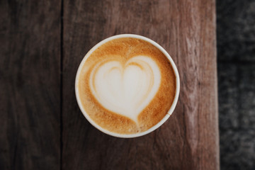 Fresh hot cappuccino coffee in a paper cup with beautiful latte art in the shape of heart on wooden background. Coffee to go.