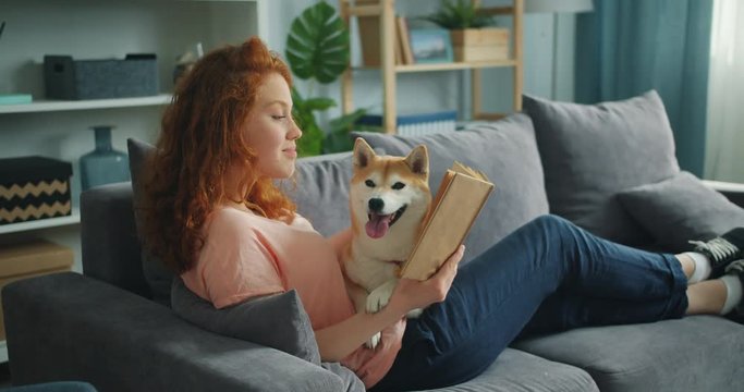 Pretty student young woman is reading book in cozy apartment smiling and petting adorable dog sitting on comfy couch at home. Animals and hobby concept.