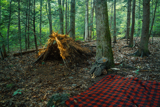 Debris hut survival shelter in the forest with Fairy Lights and a Campfire, blanket, and backpack. Bushcraft camping in the forest wilderness.