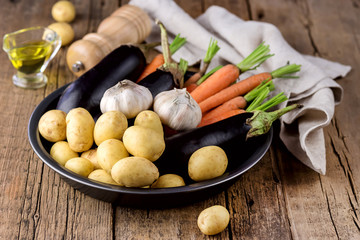 Raw Potatoes Carrot Garlic and Eggplant on Black Plate Variety of Raw Vegetables ready to Cook Fresh Natural Vegetables Organic Healthy Food Olive Oil Pepper Pot Wooden Background Horizontal