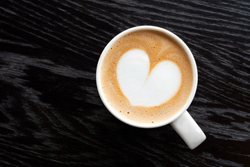 Cappuccino with heart shape foam in a white ceraamic mug isolated on dark brown wood table with grain from above. Space for text.