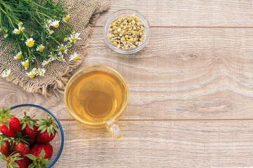 Glass cup of green tea, stawberries with white chamomile flowers on wooden background.