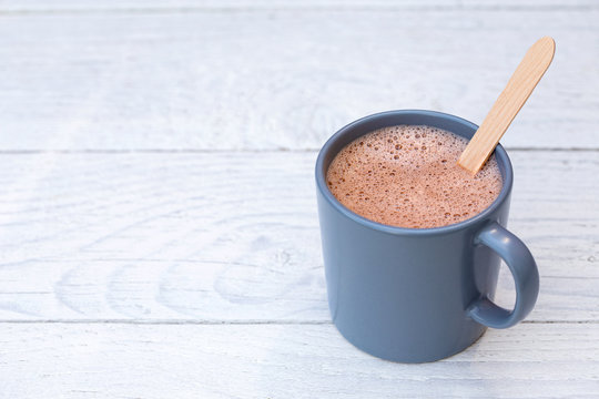 Hot chocolate in a blue-grey ceramic mug with wooden stirrer isolated on white painted wood. Space for text.