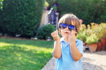 Portrait of funny little boy putting on big sunglasses with blue sky and clouds reflecting in them outdoors in garden at sunny summer day. Summer lifestyle. Curious childhood. Copy space