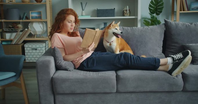 Attractive girl student is reading book and stroking lovable shiba inu puppy sitting on couch in flat enjoying literature. Hobby, youth and leisure activity concept.