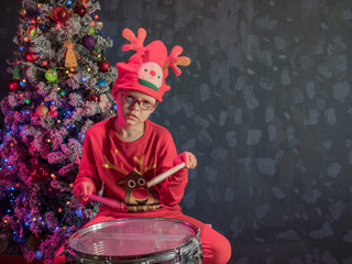 Happy Caucasian childish drummer in carnival costume Santa Claus Deer plays on a new drum set with drum sticks in his hands. Parents bought child drum as gift for Christmas. Lights on Christmas tree
