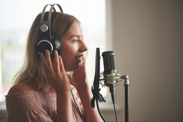 Beautiful blonde singer girl in headphones in home recording studio sings a song into a microphone,...