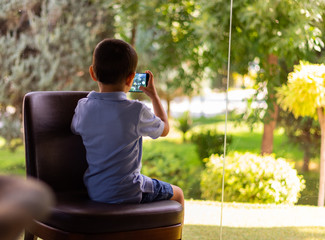 little boy in light blue t-shirt and dark blue shorts sitting on a chair and capturing an image of a beautiful green garden in a sunny day through panoramic glass window in a house on the 1st floor