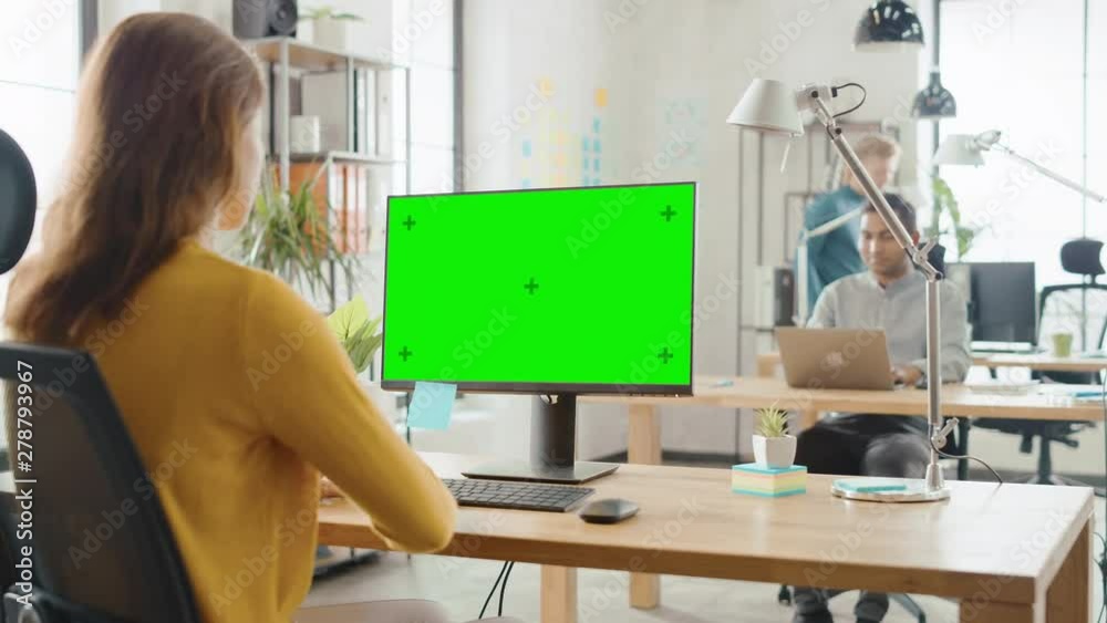 Poster over the shoulder: creative young woman sitting at her desk using desktop computer with mock-up gree