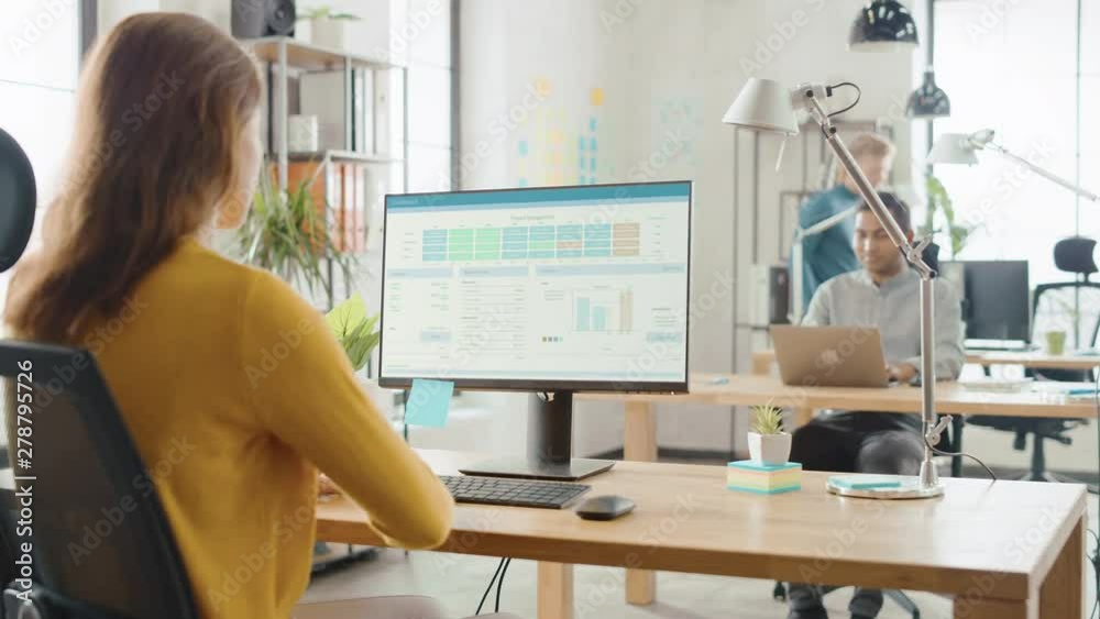 Poster Over the Shoulder: Creative Young Woman Sitting at Her Desk Using Desktop Computer with Screen Showing Project Management, Statistics and Graphs. Office with Diverse Team of Professionals Working