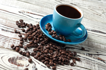 Full blue ceramic cup of black hot coffee near lot of scattered fried beans on old rustic white wooden table