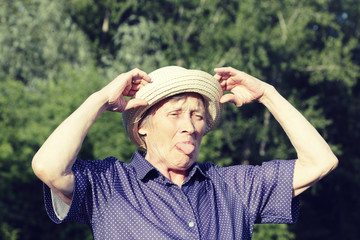 A portrait of an old woman who straightens her hat and shows her tongue, fooling around. Sunny day.