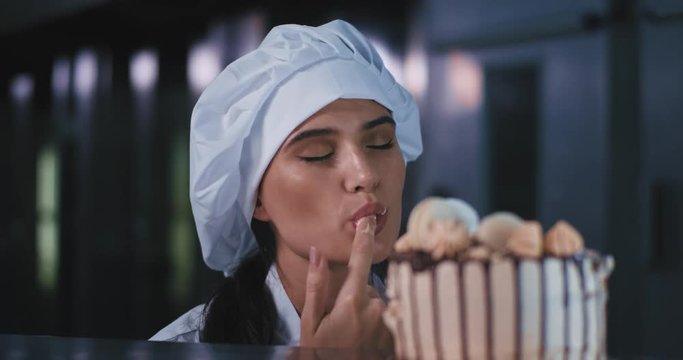 A close up shot of the sexy young baker woman with chefs attire adoring her cake and tasting it with her finger, she looks very pleased and happy