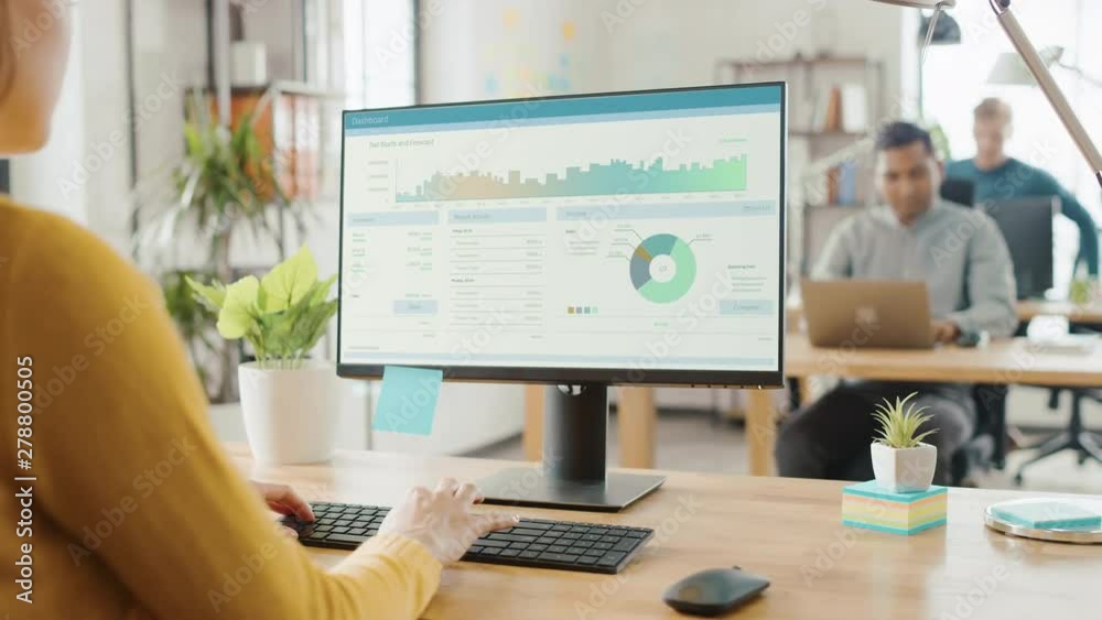 Poster Over the Shoulder: Creative Young Woman Sitting at Her Desk Using Desktop Computer with Screen Showing Project Management, Statistics and Graphs. Office with Diverse Team of Professionals Working