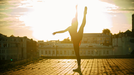 Fototapeta na wymiar Young woman ballerina training on the roof - standing in the pose showing her stretching - raised her leg up - bright sunset