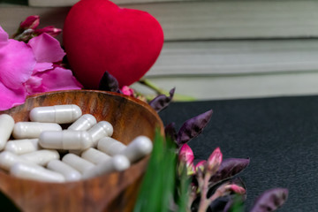 Close up image of books behind the herbal medicine capsules in a wood spoon with red heart, pink flowers and green leaf on dark background - Herbal Aurvedic Medicine for heart Concept