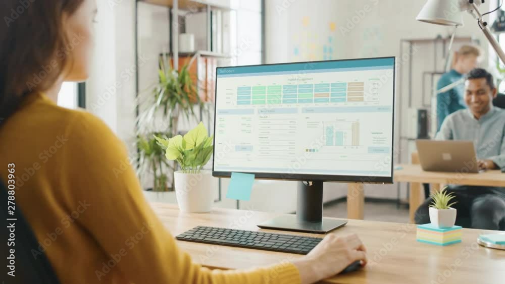 Poster Over the Shoulder: Creative Young Woman Sitting at Her Desk Using Desktop Computer with Screen Showing Project Management, Statistics and Graphs. Office with Diverse Team of Professionals Working