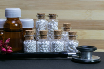 Natural Homeopathy Concept  -  A Stethoscope with homeopathic medicine bottles consisting of pills and liquid substance with pink flower bud on  wood and dark background