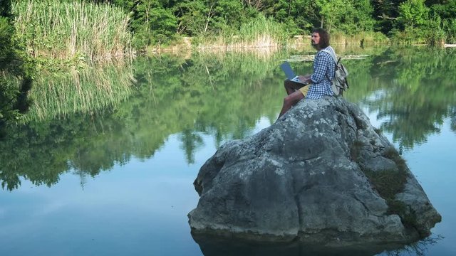 A young freelance guy sits on a stone in the middle of a lake with a laptop. Freelance concept. Work on travel. Wireless internet in the forest.
