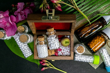 Top view image of homeopathic medicine bottles, scattered pills with wild pink flower, wild fruit in wooden old box on burlap and green leaf with dark background. Homeopathic medicine concept