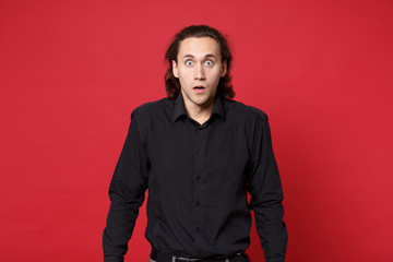 Fototapeta premium Shocked upset stylish young curly long haired man in black shirt posing isolated on red wall background studio portrait. People sincere emotions lifestyle concept. Mock up copy space. Looking camera.