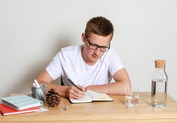 A young man in a white t-shirt sitting at his Desk, writing something in a notebook.