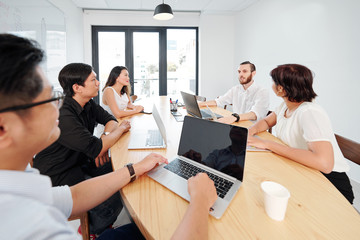 Young bearded businessman sitting at the table with laptop computer and talking while other business people listening to him during teamwork at office