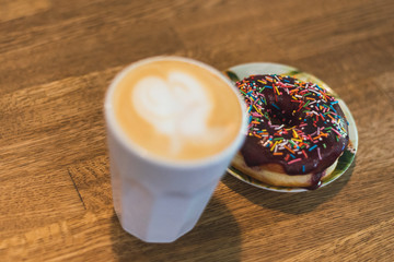 coffee with a heart made of milk and a donut on a wooden table. Coffee with a drawn heart and milk on a wooden table in a coffee shop. chocolate donut with scattering on the table next to the coffee