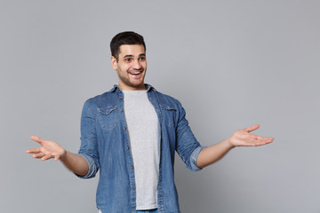 Shocked stylish unshaven young man in denim jeans shirt posing isolated on grey wall background studio portrait. People sincere emotions lifestyle concept. Mock up copy space. Spreading hands wow news