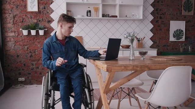 Young or adult disabled man sitting in comfort wheel chair and spending his holiday at modern flat with bright and light room inside. Male using personal computer and checking mail