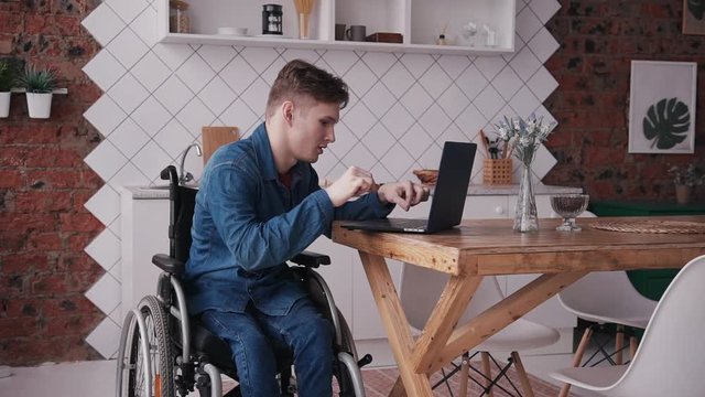 Young And Disabled Man Sitting In Special Comfort Wheelchair And Spending His Holiday At Home With Bright Light Room In Modern Interior Flat. Male Using Personal Computer Or Laptop And Working