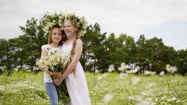 Two girl friend in wreath with flowers bouquet embracing while posing on green field. Young fashion model posing with summer flowers on outdoor photo session