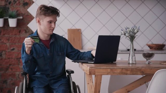 Young and disabled man sitting in special comfort wheelchair and spending money in online shopping. Male using credit card and personal laptop at home with bright cozy room with modern interior