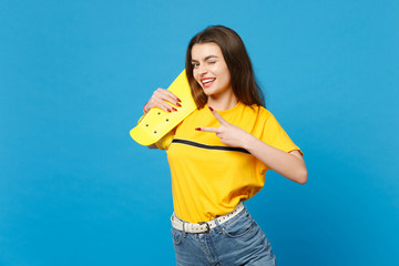 Portrait of blinking young woman in vivid casual clothes looking camera hold yellow skateboard showing victory sign isolated on blue background in studio. People lifestyle concept. Mock up copy space.