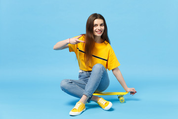 Pretty young woman in vivid casual clothes looking camera, showing victory sign sitting with yellow skateboard isolated on blue wall background in studio. People lifestyle concept. Mock up copy space.
