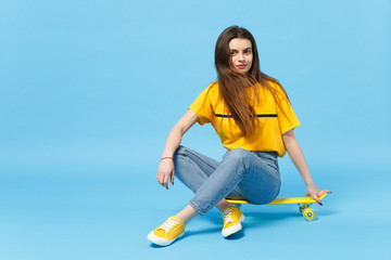 Portrait of stunning young woman in vivid casual clothes looking camera and sitting with yellow skateboard isolated on blue wall background in studio. People lifestyle concept. Mock up copy space.