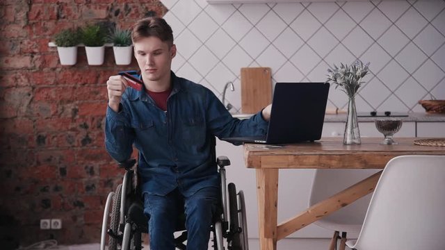 Online shopping concept. Young and disabled man sitting in comfort wheel chair and spending weekend at cozy bright room in house with modern interior. Male using bank card and pc laptop