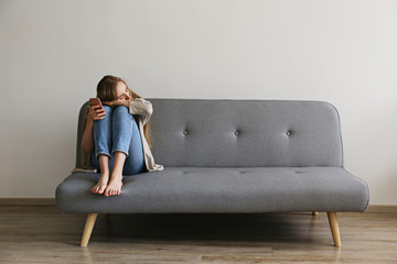Portrait of beautiful young woman with depressed facial expression sitting on grey textile couch...